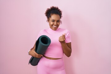 Young hispanic woman with curly hair holding yoga mat over pink background doing money gesture with...
