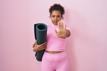 Young hispanic woman with curly hair holding yoga mat over pink background doing stop sing with...