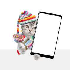 Cute kitten wearing warm hat with pompon and warm scarf holds smartphone with white blank screen in it paw above empty white banner. isolated on white background