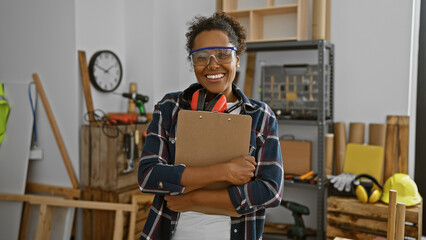 Smiling woman with safety goggles and headphones holding a clipboard in a carpentry workshop