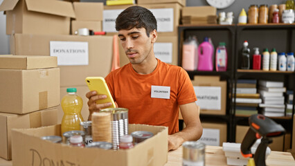 Dedicated young hispanic man volunteer earnestly checking charity products on his smartphone at...
