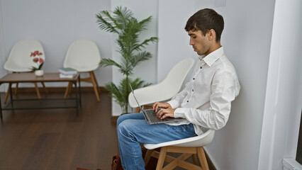 Handsome young hispanic man engrossed in serious business on laptop, sitting in chair at bustling...