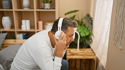 Obraz premium Handsome, grey-haired, young hispanic man comfortably sitting on the bed, relaxed in his cozy bedroom, enjoying his morning, lost in the rhythm of his favourite music, wearing headphones.