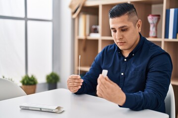 Young latin man sitting on table holding antigen test at home