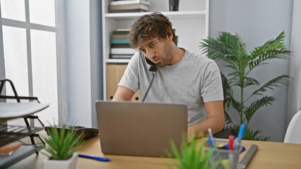 A focused man working and multitasking with a laptop and telephone in a modern office setting.