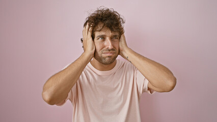 A troubled young hispanic man in a pink shirt stands against a pink wall, looking frustrated with hands on his head.