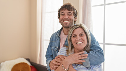 Confident mother and son share a warm, smiling hug indoors, enjoying their love-filled, casual...