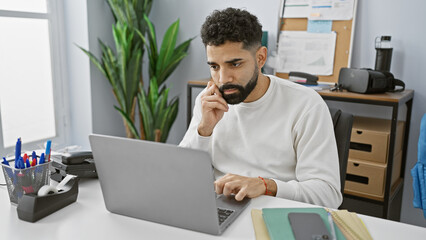 Concentrated hispanic man working in a modern office, using a laptop and wearing casual clothes.