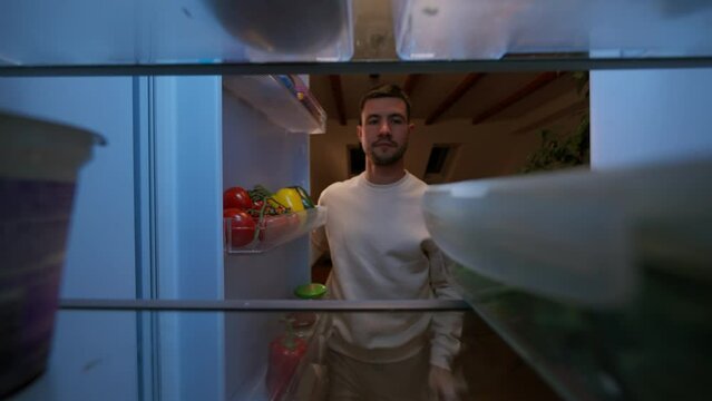 View inside kitchen fridge: handsome man opens fridge door, putting inside salad in box