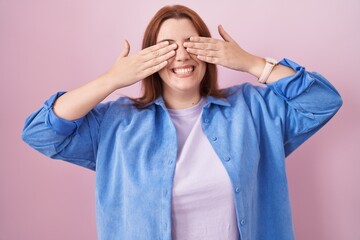 Young hispanic woman with red hair standing over pink background covering eyes with hands smiling...