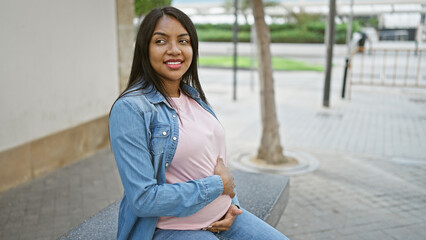 Cheerful young pregnant woman, confidently touching belly, sits carefree on a city bench, enjoying...