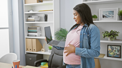 Smiling young pregnant businesswoman touching her belly, working with laptop at the office, displaying confidence and success