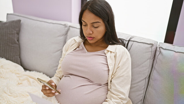 Relaxed young hispanic woman, joyfully wrapped in a blanket, sitting on her home's living room sofa, discloses her pregnancy news online using her smartphone, completely absorbed in motherhood bliss.