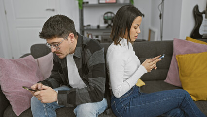 A man and woman seated back to back on a couch, engrossed in smartphones in a modern living room.