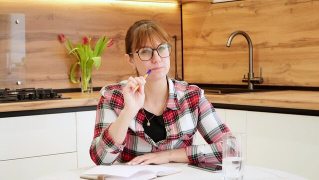 Portrait of a thoughtful and sensible woman with glasses writing a list of household products. A lady with a notebook and pen sits at the kitchen table and decides what to buy and cook for dinner.