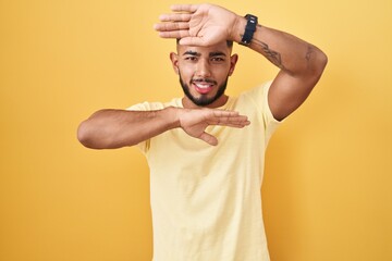 Young hispanic man standing over yellow background smiling cheerful playing peek a boo with hands...