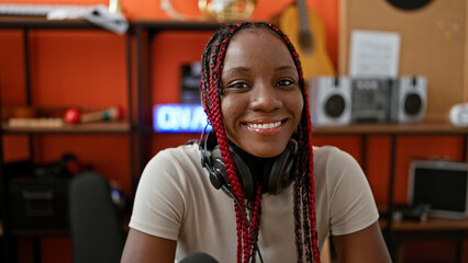Smiling african american female musician wearing headphones, sitting at table in music studio,...