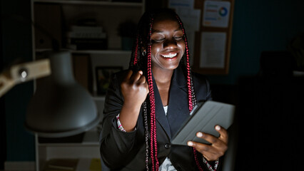 Confident african american woman worker celebrating business success with touchpad at office, a...