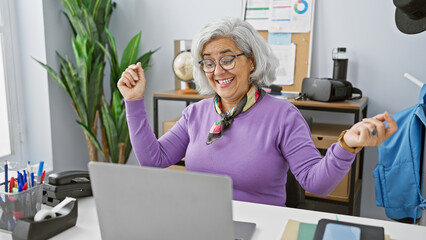 A joyful middle-aged woman with grey hair celebrating success in a modern office setting.