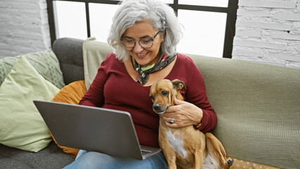 A smiling woman with grey hair cuddles her dog while using a laptop in a cozy living room interior.