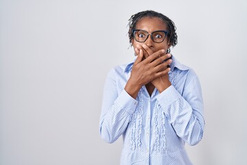 African woman with dreadlocks standing over white background wearing glasses shocked covering mouth...
