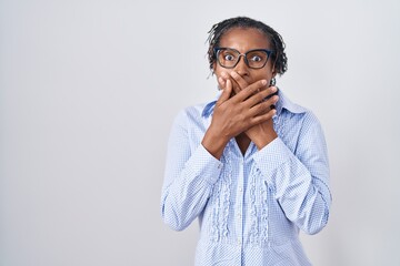 African woman with dreadlocks standing over white background wearing glasses shocked covering mouth with hands for mistake. secret concept.