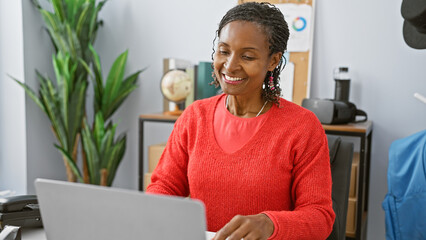 A smiling african woman in a bright red sweater working on a laptop in a modern office setting.