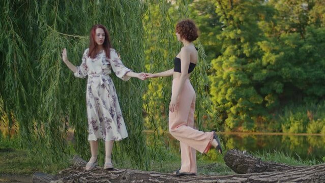 Two women balancing and holding hands on a fallen tree trunk in a park. Friendship and trust concept. Design for outdoor activities, teamwork building, and nature connection themes. Full-length shot