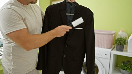 A man in a white shirt lint-rolling a suit jacket in a bright laundry room.
