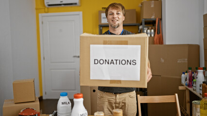 Smiling young man holding a donations box in the center of a cluttered room, symbolizing charity...