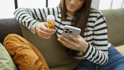 Hispanic young woman holding medication and smartphone indoors on sofa, looking at phone