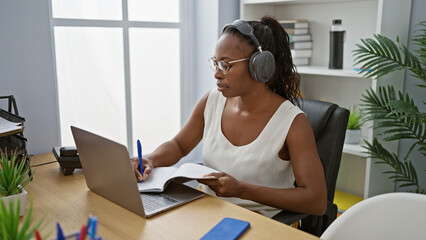 African american woman wearing headphones taking notes at her office desk indoors