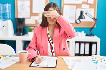 Young hispanic woman working at the office wearing glasses covering eyes with hand, looking serious...