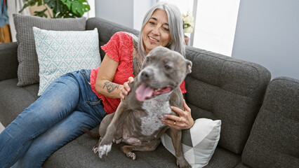 Joyful middle age woman sitting on the sofa at home, playing with her beloved grey-haired dog,...