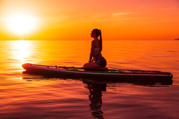 Slim woman sitting on stand up paddle board at sea with bright sunset or sunrise. Woman relax on...