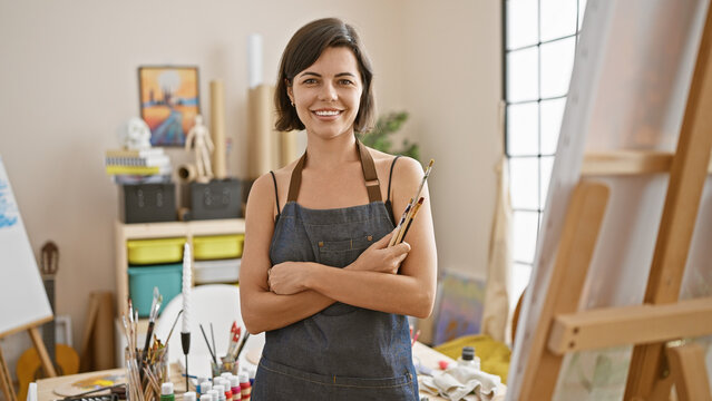 Confident young hispanic artist, a beautiful woman standing in the art studio with crossed arms, her smile revealing the joy of creativity as she holds her trusted paintbrushes.
