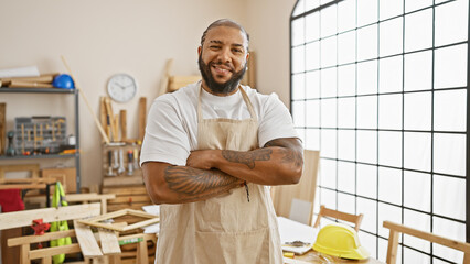 Smiling bearded man with arms crossed in a sunlit carpentry workshop, wearing an apron indicative of work.