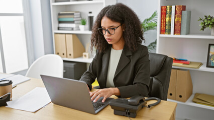 Striking curly-haired young hispanic woman, relaxed yet serious, elegantly working online on laptop...