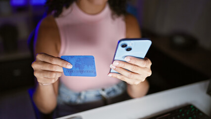 Hispanic woman streamer's hands typing, texting, paying online with credit card amidst the dark room's gaming console, streaming game on smartphone while sitting at home office table