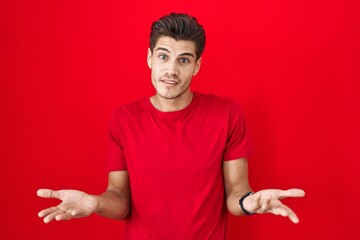 Young hispanic man standing over red background clueless and confused with open arms, no idea...