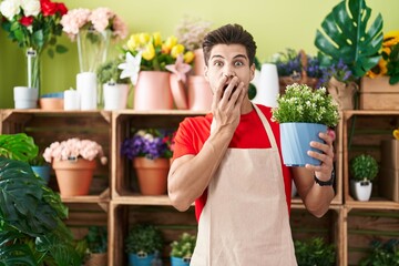 Young hispanic man working at florist shop holding plant covering mouth with hand, shocked and...