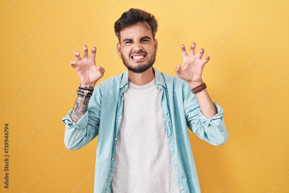 Poster Young hispanic man with tattoos standing over yellow background smiling funny doing claw gesture as cat, aggressive and sexy expression