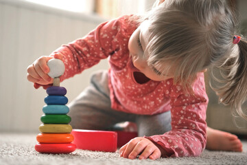 Kid girl play toys at home, kindergarten or nursery. Baby child playing playthings cubes and wood puzzle, pyramid sitting on the floor. Happy preschool education indoor. 3 years old little toddler.
