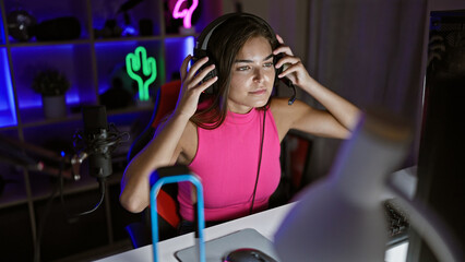Fototapeta na wymiar Attractive young hispanic female streamer, caught in the cyber night, fully concentrated and wearing headphones while playing a high-stakes video game in her dark gaming room at home