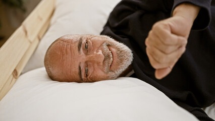 A smiling senior hispanic man with a beard and grey hair resting on a white pillow, giving a thumbs-up in a bedroom.
