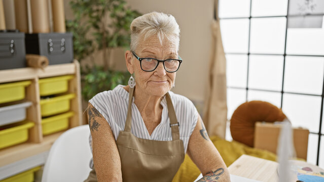 Grey-haired senior woman artist, with a serious face, paints intensely while sitting at a table in her studio