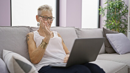 Confident grey-haired senior woman, smiling and enjoying restful moment on sofa at home, engaging with technology using laptop for relaxing video call