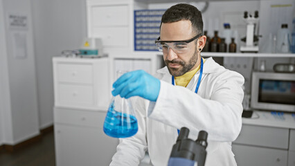 Naklejka na ściany i meble A hispanic man in a lab coat examines a chemical in a flask at an indoor laboratory, portraying a professional scientific environment.