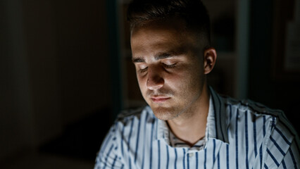 A young hispanic man with closed eyes appears to be dozing off in a dimly lit office room.