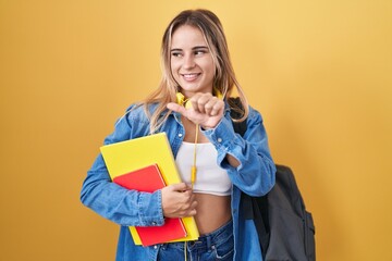 Young blonde woman wearing student backpack and holding books pointing to the back behind with hand and thumbs up, smiling confident